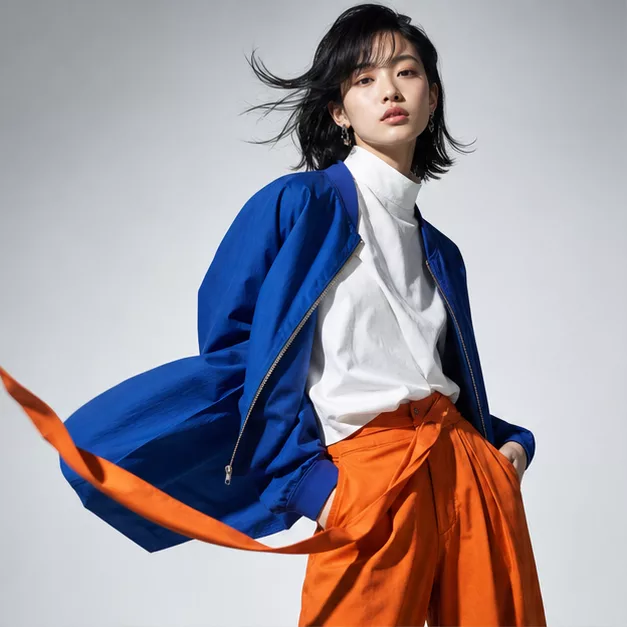 Model in a cobalt windbreaker and signal-orange trousers, hair in motion against a chalk backdrop.