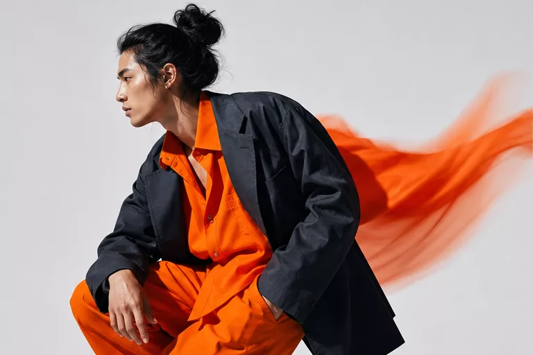 Seated model in an orange shirt and charcoal blazer with an orange fabric ribbon streaking behind.